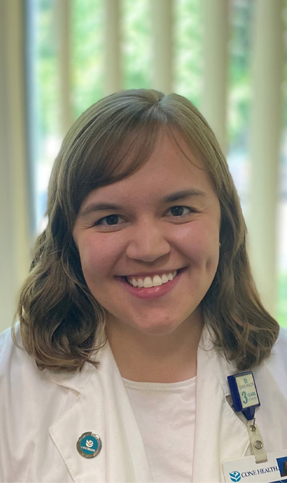 Pharmacist Catie Harper in a white lab coat in the office with blinds.