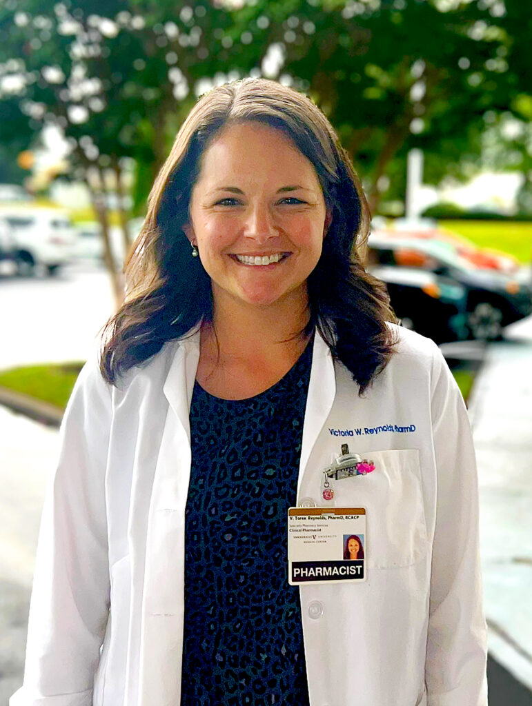 Toree Reynolds in a white lab coat, smiles at the camera. She is standing in a parking lot with parked cars behind her.