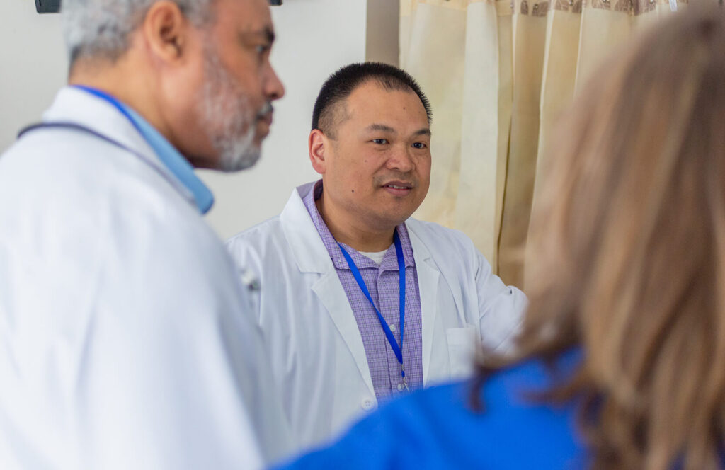 Two pharmacists in white lab coats engage in a discussion in a clinical setting, with a third colleague partially visible in the foreground. The pharmacist in the center, wearing a purple plaid shirt and blue lanyard, listens and speaks attentively during what appears to be a team consultation near a privacy curtain.
